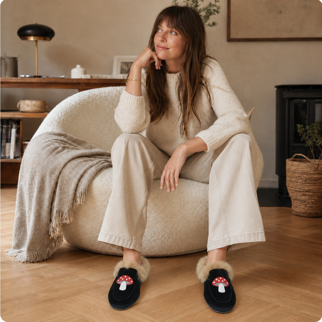 Mules fourrées à motif champignon en velours noir portées par une femme assise dans un fauteuil bouclé, pull crème et pantalon beige