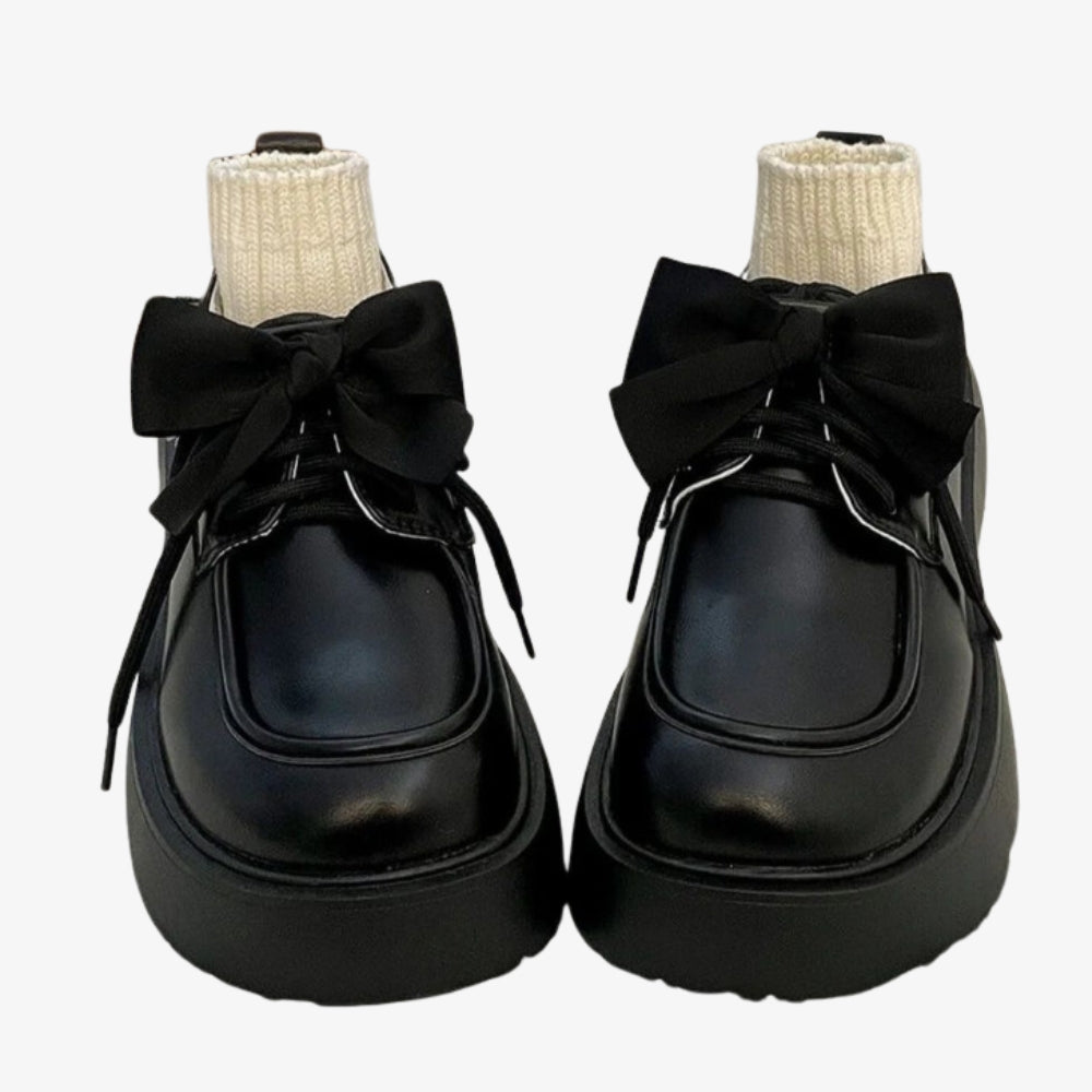 Une paire de mocassins en cuir lisse noir posée sur un lit blanc, un grand noeud décore le dessus et des petites chaussettes beiges remplissent la chaussure.