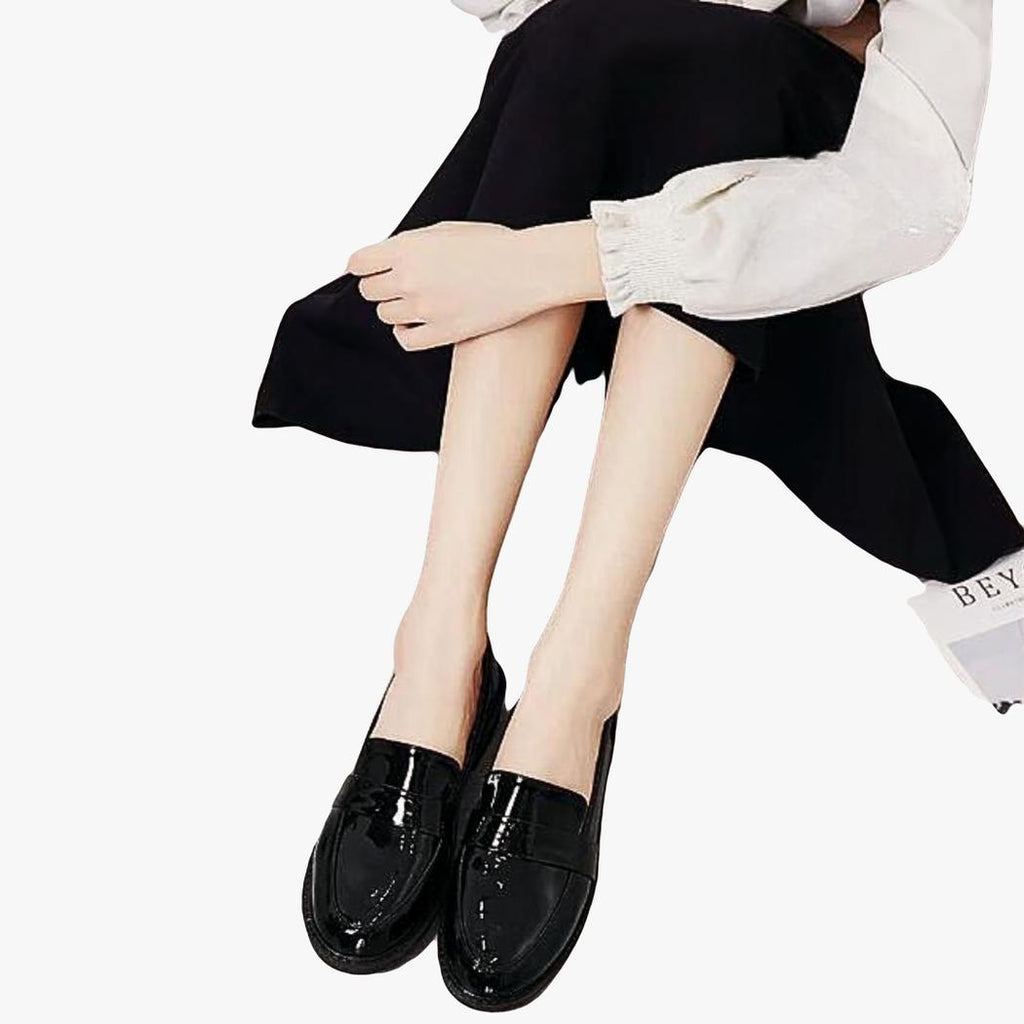 on voit une femme assise par terre on ne voit pas le haut du corps, elle porte une paire de mocassins noirs à talons carrés dans le style preppy