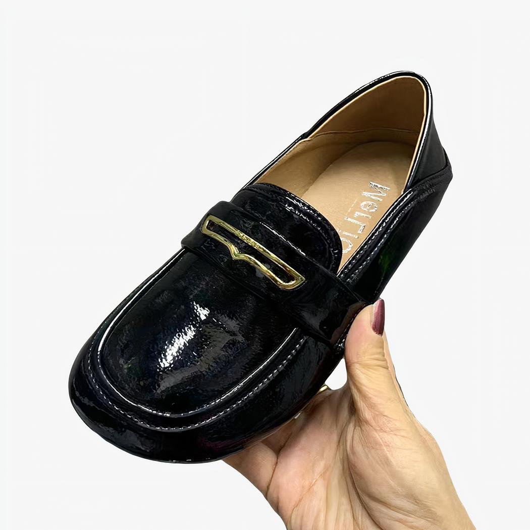 mocassin noir en cuir verni synthétique, porté à la main sur fond de mur gris