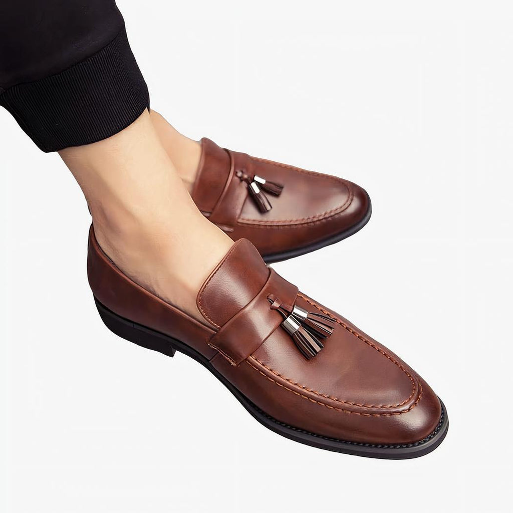 Des mocassins en cuir marron à bout pointus et glands décoratifs aux pieds d'un homme portant un pantalon noir et jambes croisées.