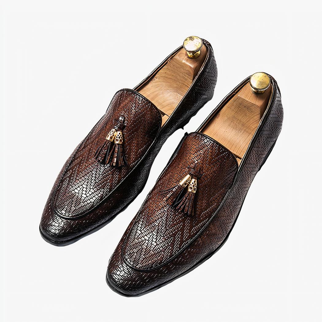Des mocassins en cuir avec des motifs de zigzag et des glands décoratifs en cuir avec un empiècement en métal doré, des bouts pointus et foncés, sur un sol quadrillé blanc et noir.