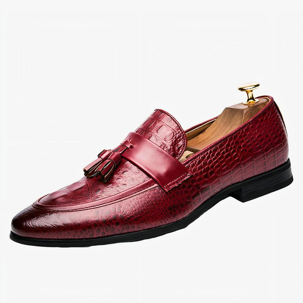 Un mocassin en cuir rouge crocodile avec gland décoratif et bout pointu et foncé sur un fond blanc.