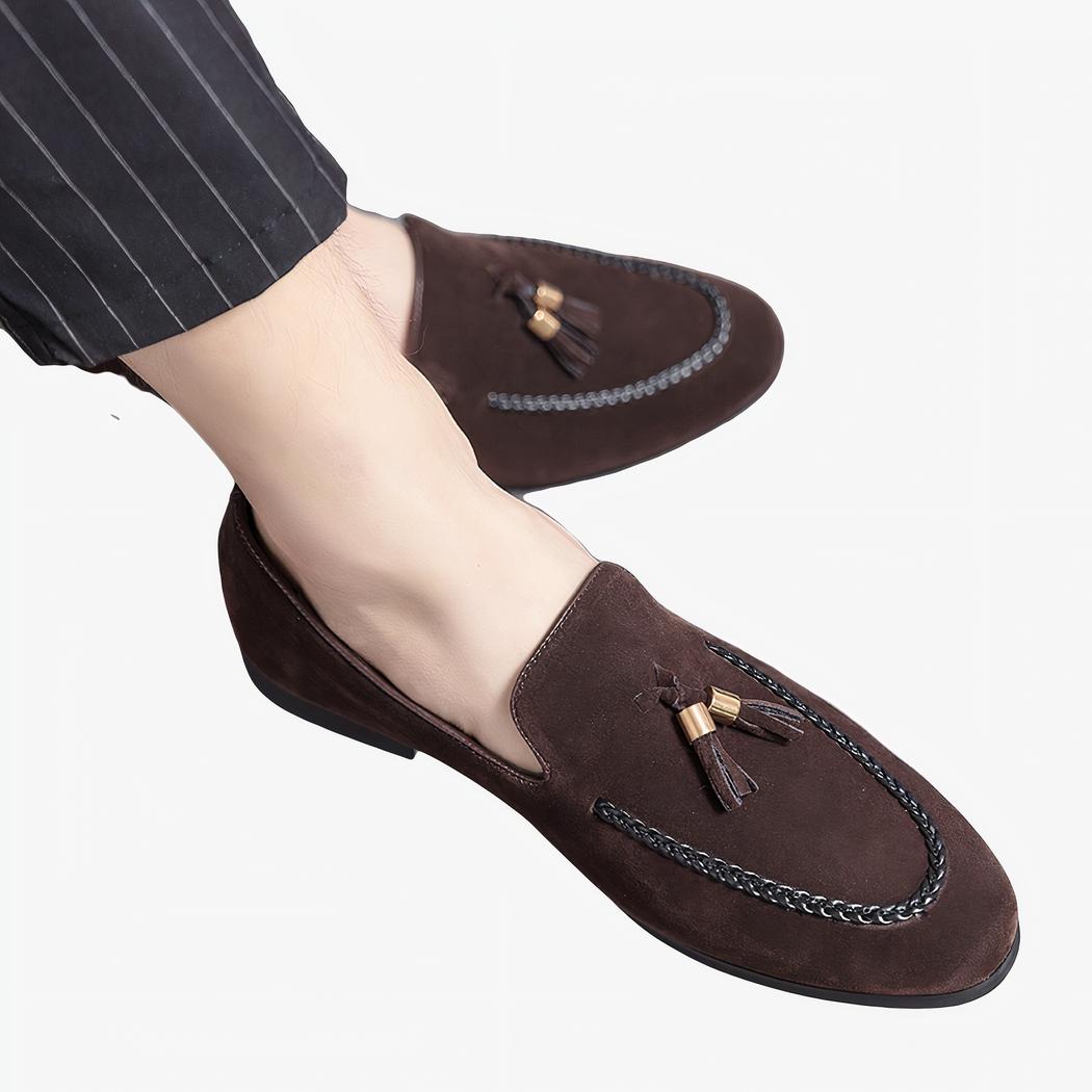 Des mocassins en daim avec glands décoratifs et une torsade en cuir sur la couture, aux pieds d'un homme aux jambes croisées portant un pantalon de costume noir à rayures fines blanches.