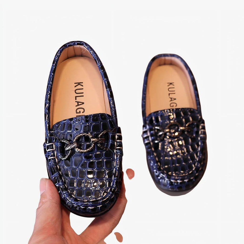 mocassin en faux cuir à motif croco pour bébé avec un anneau sur le dessus, ces chaussures sont présenter dans la main d'une femme