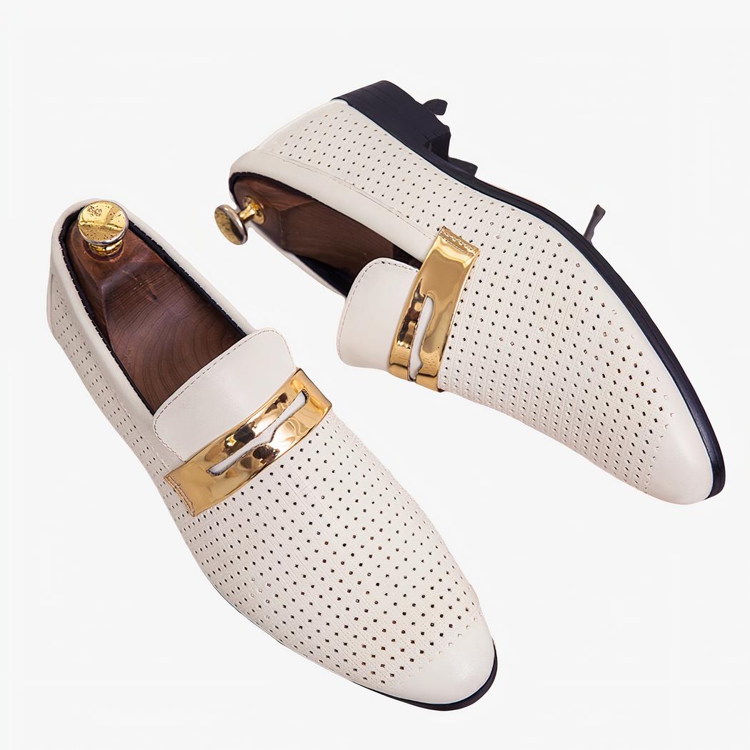 Sur une boite en bois clair, une paire de mocassins blancs de forme allongée ajourés de petits trous avec une lanière doré sur le dessus. Il y a des embauchoirs en bois avec un bouton doré au niveau du talon à l'intérieur des chaussures.