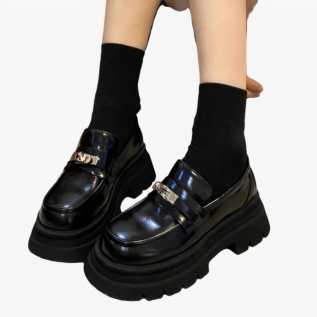 Des mocassins noirs en cuir lisse brillant avec une épaisse semelle aux pieds d'une femme assise sur le sol et portant des chaussettes courtes noires.