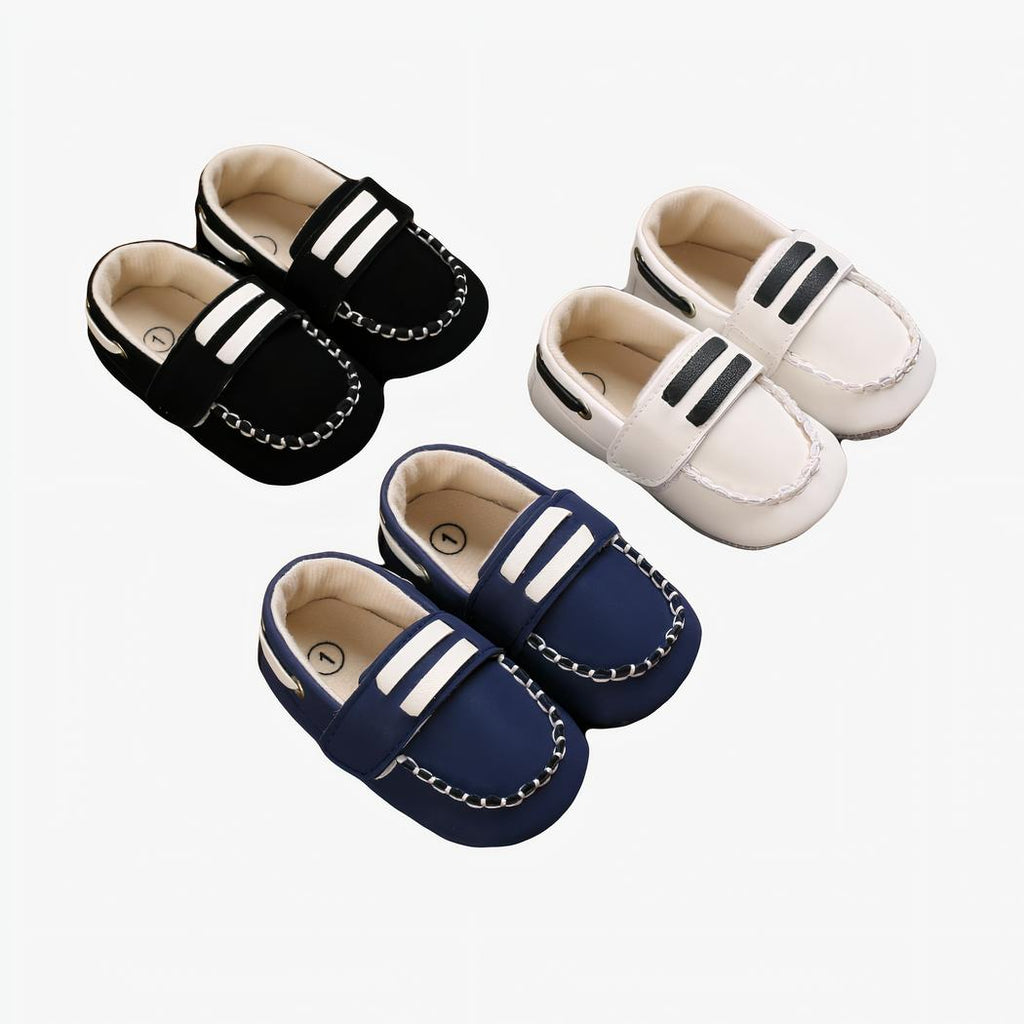 Sur parquet marron , trois paires de mocassins bateau pour bébé avec surpiqures, et deux barres blanches sur la languette du dessus. On voit une paire blanche, bleu et noire