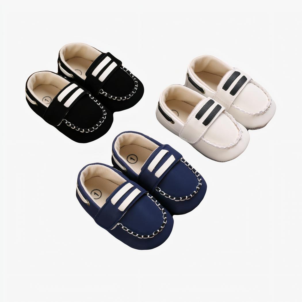 Sur parquet marron , trois paires de mocassins bateau pour bébé avec surpiqures, et deux barres blanches sur la languette du dessus. On voit une paire blanche, bleu et noire
