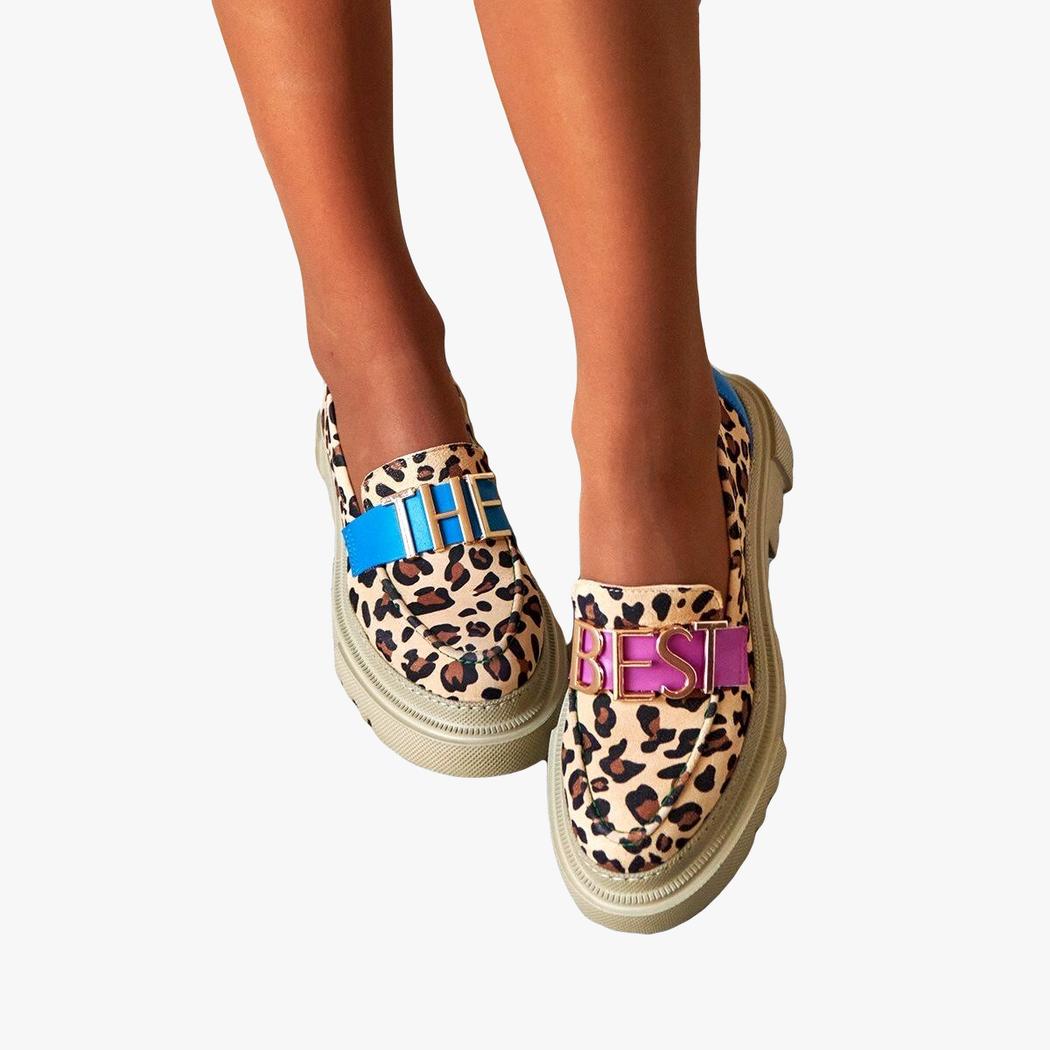 Sur un sol extérieur, une paire de mocassins portées de face en beige avec semelle plateforme épaisse, avec imprimé léopard avec une sangle bleue dessus turquoise avec lettre dorées "THE" sur une chaussure et une sangle rose avec "BEST" sur l'autre
