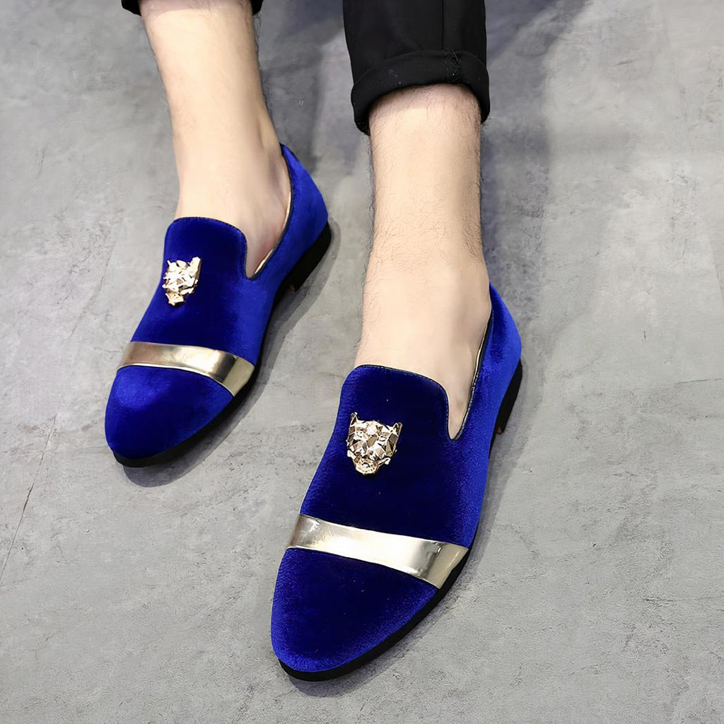 Sur sol gris, des mocassins en velours bleus avec lanière dorée sur le devant et écusson doré en tête de tigre sur le dessus. Il sont portés pieds nus