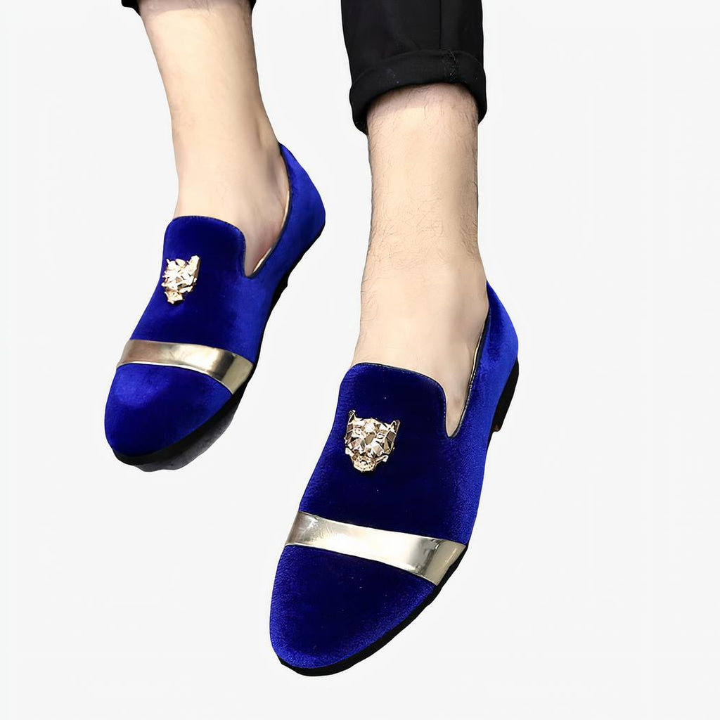 Paire de mocassins en velours bleu brillant avec lanière dorée sur l'avant et écusson en tête de tigre en relief sur le dessus. Ils sont portés avec un pantalon noir sur un sol gris