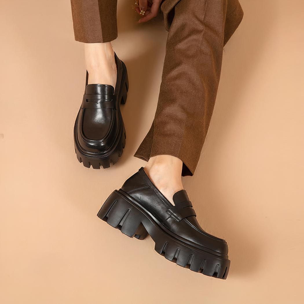 Photo d'une paire de mocassins noire portée par une femme avec un pantalon marron sur fond rose