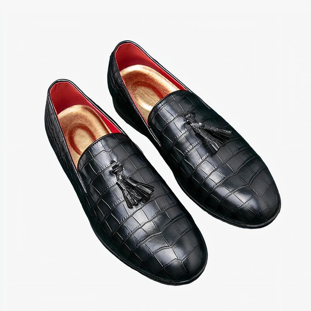 Paire de mocassins noirs à glands posés sur le sol bleu avec des lunettes rondes posées à côté.