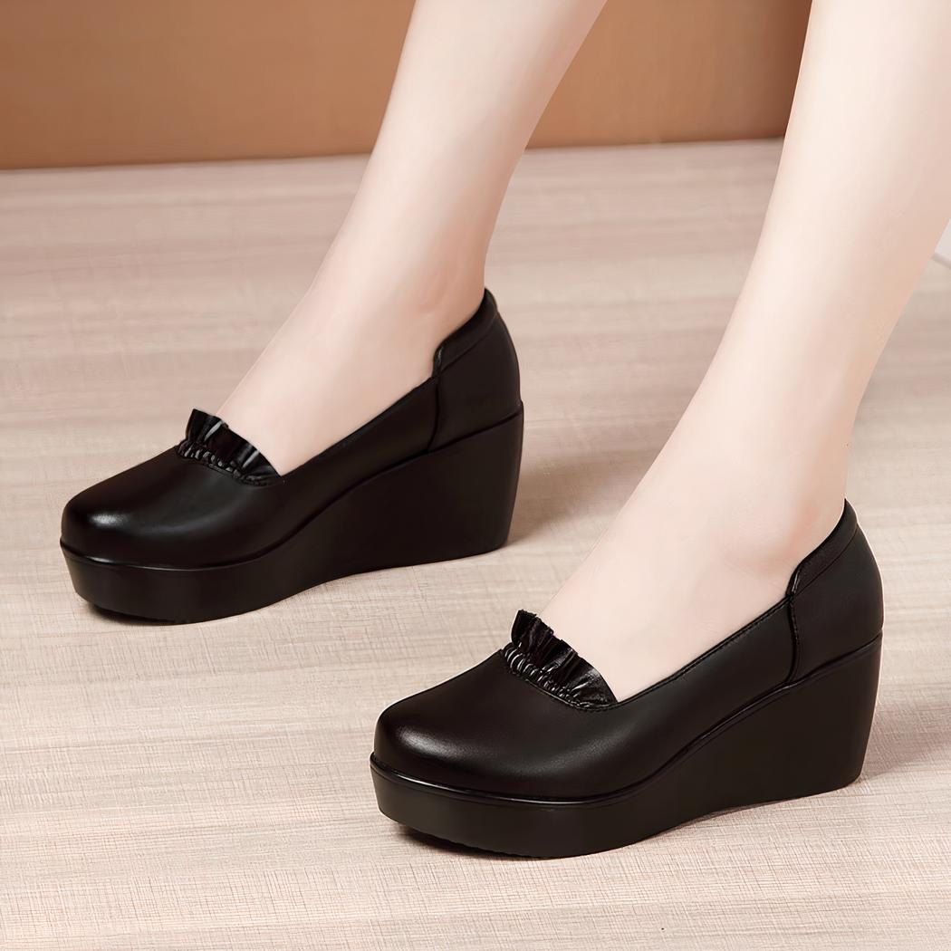 Photo d'une paire de mocassins noire compensée portée par une femme sur un parquet et avec un mur marron en fond
