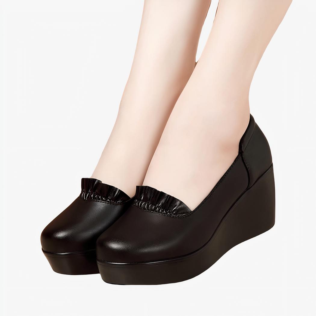 Photo d'une paire de mocassins noire compensée portée par une femme sur un parquet et avec un mur marron en fond