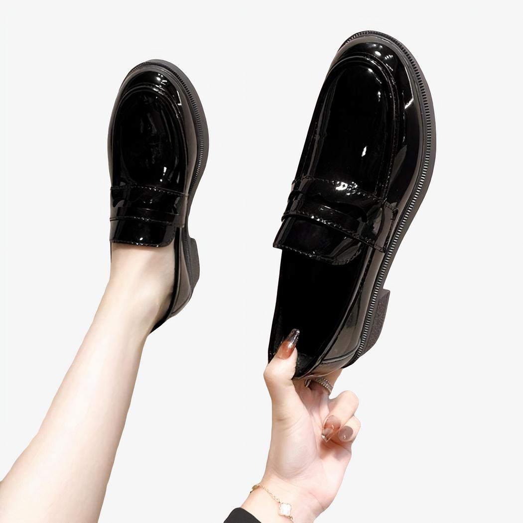 Mocassins noirs en cuir vernis, un porté l'autre tenu à la main, sur fond gris