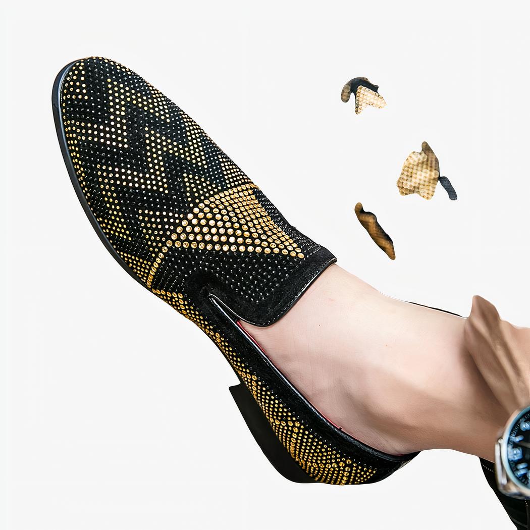 Portée pieds nus, une paire de mocassins en noir avec broderies en forme géométriques en strass doré. Un pied est posé au sol sur un parquet marron.