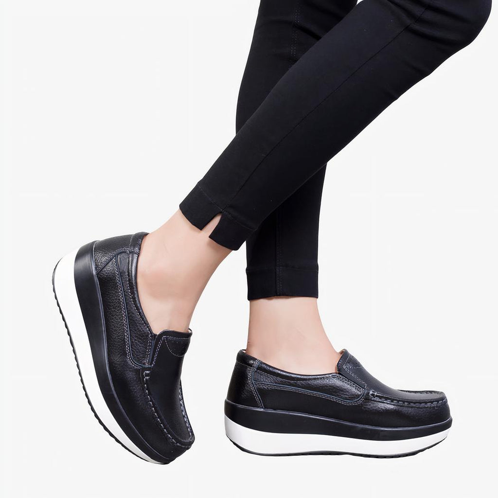 Photo de mocassins orthopédiques à plateforme noirs avec une semelle blanche, portée par des jambes de femme en pantalon noir.
