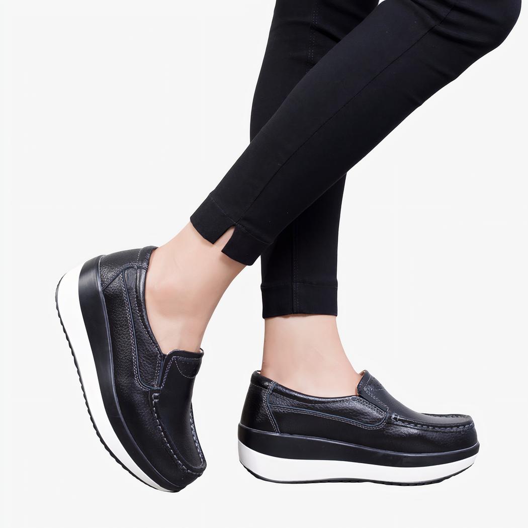 Photo de mocassins orthopédiques à plateforme noirs avec une semelle blanche, portée par des jambes de femme en pantalon noir.