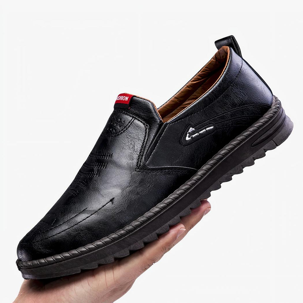 Mocassin noir avec une semelle épaisse et tenu dans une main. Fond gris.
