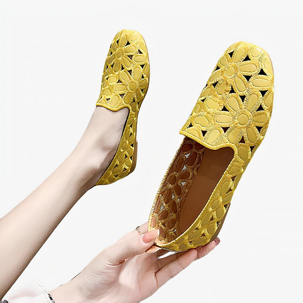Paire de mocassins plats souples jaunes ajourés avec motifs fleurs. Une chaussure est dans un pied appuyé contre un mur gris et l'autre est tenue dans une main