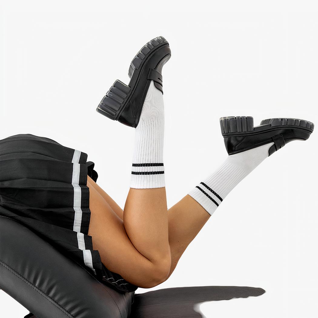 Des mocassins noir à talon et semelle épaisse noir et semi translucide aux pieds d'une femme portant une jupe plissée et des chaussettes blanches dans une position allongée les pieds en l'air.