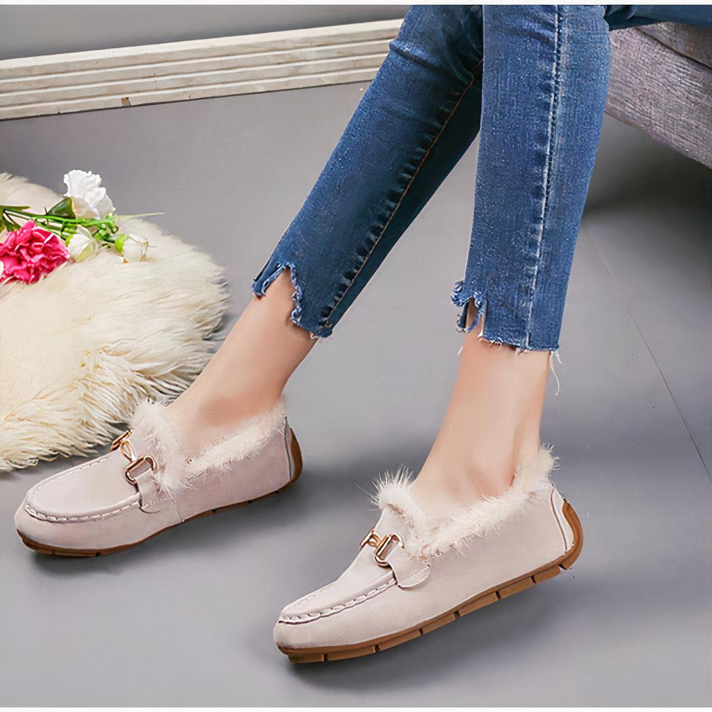 Paire de mocassins hiver suède beiges avec fourrure apparente portée avec un jean et le même modèle en noir posé à coté. Le sol est gris et il y a un bout de tapis en fourrure beige dessus.