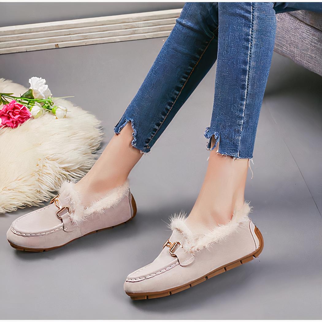 Paire de mocassins hiver suède beiges avec fourrure apparente portée avec un jean et le même modèle en noir posé à coté. Le sol est gris et il y a un bout de tapis en fourrure beige dessus.