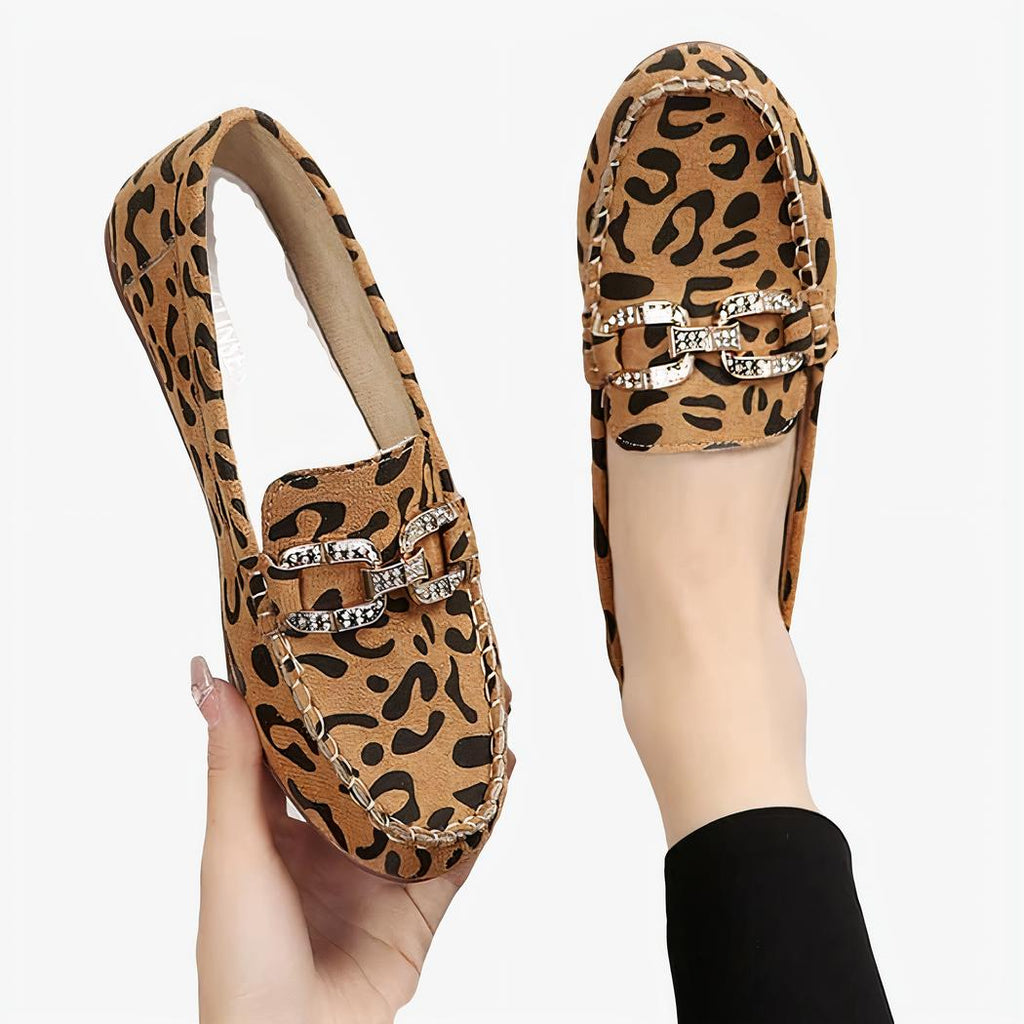 Une paire de mocassins arrondis avec imprimé léopard marron et anneaux strassés argentés, une chaussure présentée au pied avec un collant noir et l'autre tenue dans une main