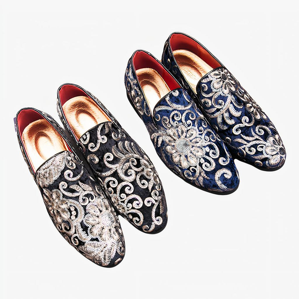 Deux paires de mocassins noirs et bleus en velours brillant avec broderie fleurie et sequins avec une semelle intérieure dorée. Ils sont posés sur une surface en bois marron à coté d'un magazine de vêtements homme