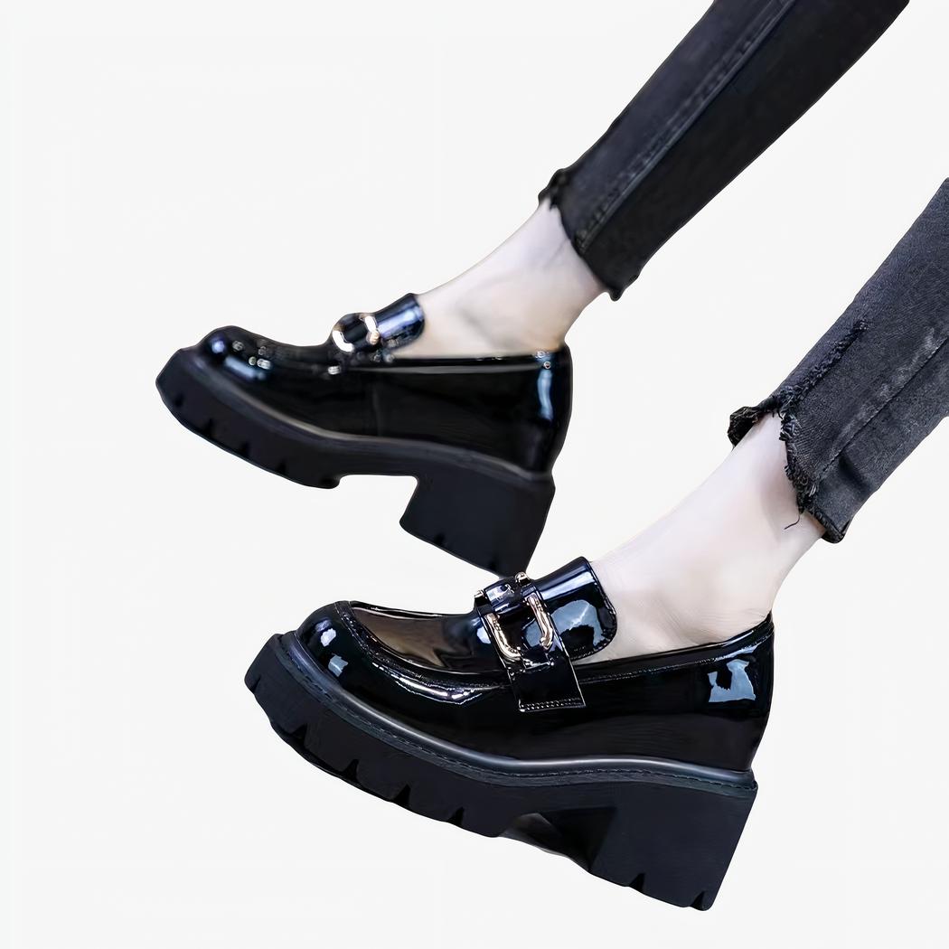 Des mocassins noirs vernis à talon et semelle épaisse noire aux pieds d'une femme se tenant debout et portant un jean noir slim.