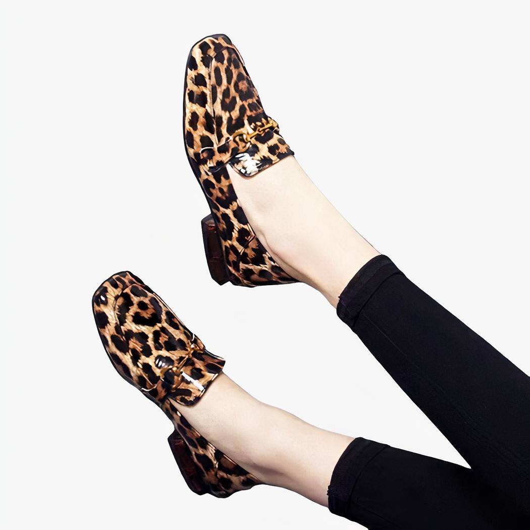 Sur fond gris, paire de jambes avec legging noirs et mocassins brillants avec talons carrés imprimé léopard