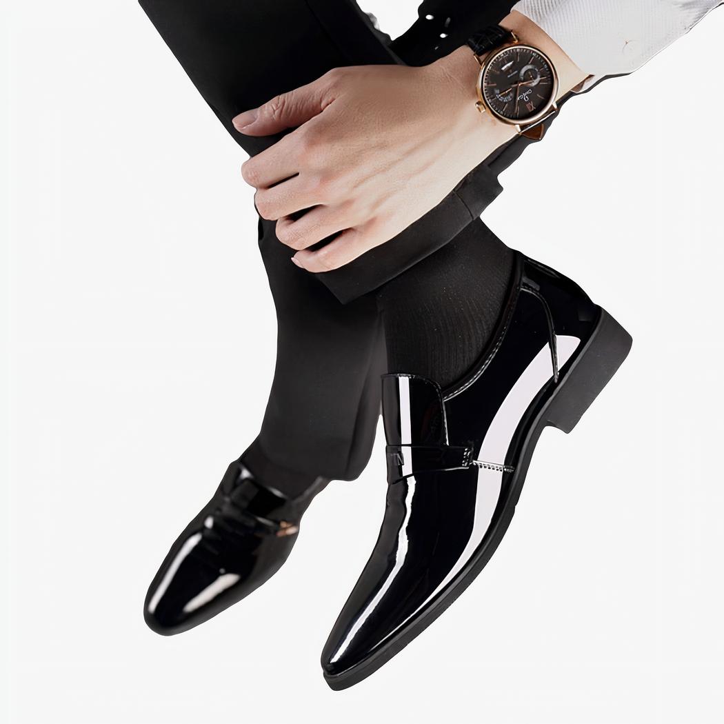 Pieds avec des mocassins noirs en cuir vernis. Jambe croisée ave une montre au poignet.