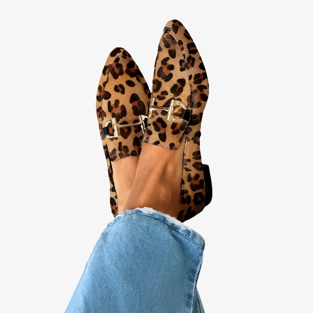 Portée avec un jean et chevilles apparentes, on voit de profil une paire de mocassins imprimé léopard marron avec petit talon imitation bois et une chaine dorée en décoration sur le dessus. Il y a une chaine de cheville dorée sur un pied.