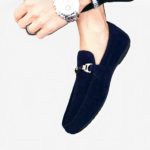 Pieds d'une personne portant des mocassins bleus avec une boucle dorée sur le devant et portant une montre au poignet