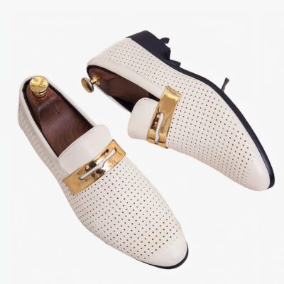 Sur une boite en bois clair, une paire de mocassins blancs de forme allongée ajourés de petits trous avec une lanière doré sur le dessus. Il y a des embauchoirs en bois avec un bouton doré au niveau du talon à l'intérieur des chaussures.