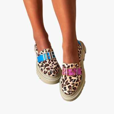 Sur un sol extérieur, une paire de mocassins portées de face en beige avec semelle plateforme épaisse, avec imprimé léopard avec une sangle bleue dessus turquoise avec lettre dorées "THE" sur une chaussure et une sangle rose avec "BEST" sur l'autre