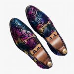 Sur une surface quadrillée avec inscriptions en noir et blanc, une paire de mocassins de soirée avec arabesques multicolores et mors argentés. Il y a des embauchoirs en bois avec un bouton doré à l'intérieur des chaussures.