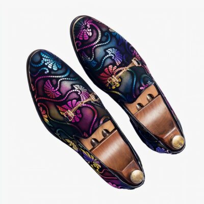 Sur une surface quadrillée avec inscriptions en noir et blanc, une paire de mocassins de soirée avec arabesques multicolores et mors argentés. Il y a des embauchoirs en bois avec un bouton doré à l'intérieur des chaussures.