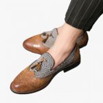 Sur sol gris, une paire de mocassins habillés pour hommes bi matière en cuir synthétique marron marbré avec dessin en surpiqure et en toile rayé grise avec gland sur le dessus.