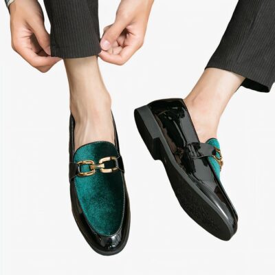Paire de mocassins pour hommes vernis noir sur le tour et en velours vert brillant avec mors doré sur le dessus. Ils sont portés avec un pantalon noir sur un sol en parquet marron