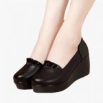 Photo d'une paire de mocassins noire compensée portée par une femme sur un parquet et avec un mur marron en fond