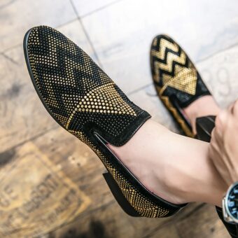 Portée pieds nus, une paire de mocassins en noir avec broderies en forme géométriques en strass doré. Un pied est posé au sol sur un parquet marron.