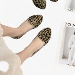 Paire de mocassins simple avec imprimé léopard vue du haut porté avec une jupe blanche et pieds nus. Le sol est en moquette beige et il y a un magazine blanc à côté