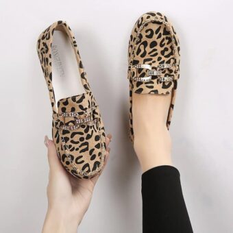 Paire de mocassins en motif léopard avec surpiqures et anneaux en strass argentés sur le dessus. Une chaussure dans un pied et l'autre tenue dans une main.