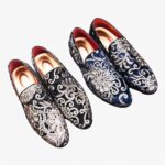 Deux paires de mocassins noirs et bleus en velours brillant avec broderie fleurie et sequins avec une semelle intérieure dorée. Ils sont posés sur une surface en bois marron à coté d'un magazine de vêtements homme