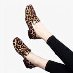 Sur fond gris, paire de jambes avec legging noirs et mocassins brillants avec talons carrés imprimé léopard