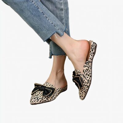 Sur un sol moquette gris, paire de mocassins en mules pointues avec nœud noir sur le dessus, un imprimé léopard orné de rivets dorés. La paire est porté avec un jean bleu court et déchiré, les chevilles sont nues.