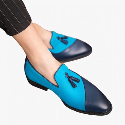 Des mocassins en cuir et textile bleu foncé et clair qui contraste avec des glands décoratifs sur les pieds d'un homme aux jambes croisées portant un pantalon de costume noir à rayures fines blanches.